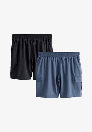 Man staat met zijn handen achter zijn rug, draagt een wit T-shirt, zwarte korte broek, witte sokken en witte sportschoenen tegen een witte achtergrond.