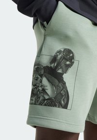 Zelené šortky s černobílým náčrtem Mandaloriana, jak drží Grogu, na levém stehně, nošené s černým tričkem.