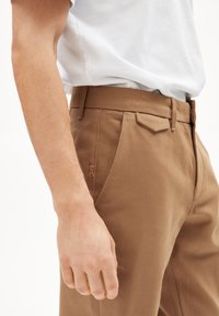 Pantalons en coton marron avec une texture lisse, poches frontales plates et coupe slim. Présente un détail de logo discret sur la poche.