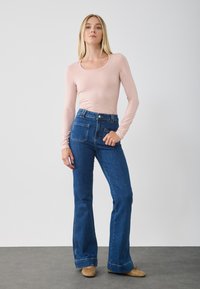 Helles rosa Langarmoberteil mit rundem Halsausschnitt, kombiniert mit dunkelblauen Schlagjeans, die über aufgesetzte Taschen und einen Knopfverschluss verfügen.