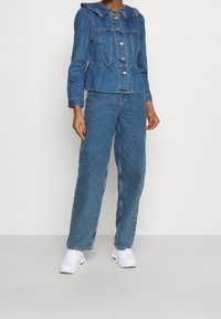 Denim jacket with a frilled collar and button front, paired with wide-leg denim pants. Both items are in a medium blue hue. White sneakers.