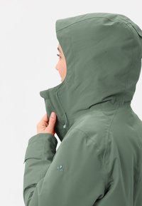 Olijfgroene jas met een capuchon, voorzien van een hoge kraag, verstelbare sluiting en subtiele logodetail op de mouw. Glad, mat textuur.