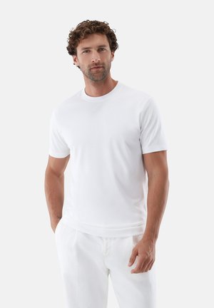 Mann mit lockigem Haar steht, trägt ein schlichtes weißes T-Shirt mit kurzen Ärmeln und weiße Hosen, vor einem hellgrauen Hintergrund.