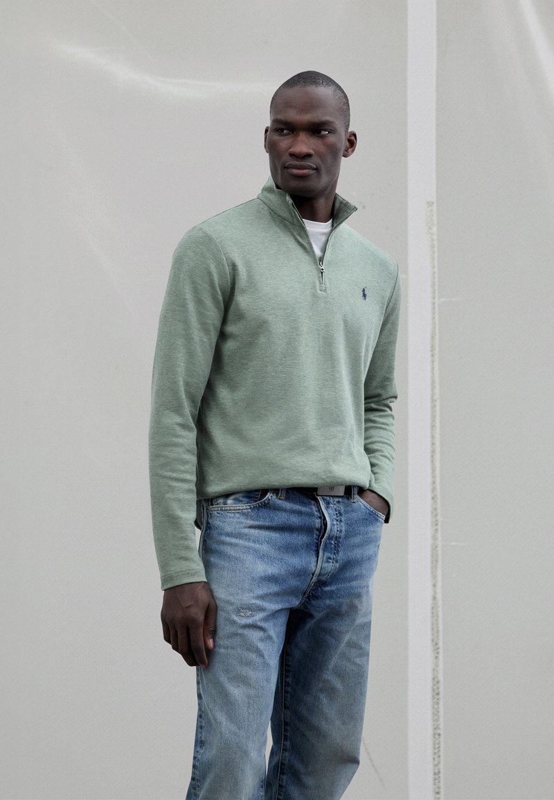 Zöld cipzáras pulóver bordázott gallérral, fehér ing fölött viselve, világoskék farmernadrággal párosítva. Kis logó található a mellrészen.