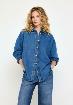 Kvinde med krøllet blondt hår iført oversized blå denimskjorte og jeans med hænderne i lommerne mod en ensfarvet hvid baggrund.