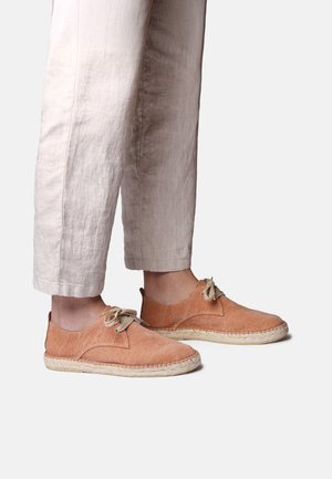 Beine in hellbeiger Hose und orangefarbenen, aus Stoff gefertigten Schnür- Espadrilles mit gewebten Sohlen vor weißem Hintergrund.