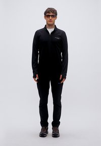 Μαύρο πουκάμισο με μακρύ μανίκι και φερμουάρ και παντελόνι από ελαφρύ ύφασμα· συνδυασμένα με γκρι και μαύρα παπούτσια· το μοντέλο στέκεται μπροστά σε ένα απλό φόντο.