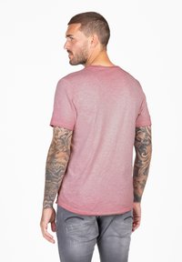 Korte mouwen, gemêleerde roze t-shirt met een ongelijkmatige zoom en ronde halslijn. Gekocht met lichtgrijze jeans, waarop gedetailleerde armtatoeages zichtbaar zijn.