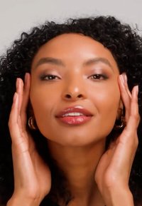 Femme aux cheveux bouclés touchant les côtés de son visage, portant des boucles d'oreilles créoles et un maquillage naturel avec des lèvres brillantes sur un fond uni.