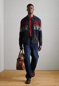 Niebieski cardigan z czerwonymi i zielonymi wzorami geometrycznymi, bordowa koszula, niebieskie dżinsy, brązowa skórzana torba i ciemno brązowe buty na drewnianej podłodze.