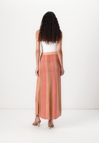 Jupe maxi avec une texture côtelée en rayures rose, pêche et or. Elle possède une taille élastique, une fente sur le côté, et est portée avec des sandales beige à lanières.