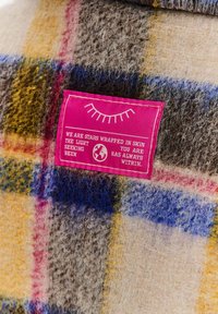 Vlněná tkanina v kostkovaném vzoru kombinující šedou, žlutou, modrou a červenou barvu se růžovou etiketou s bílým textem a grafickým designem.