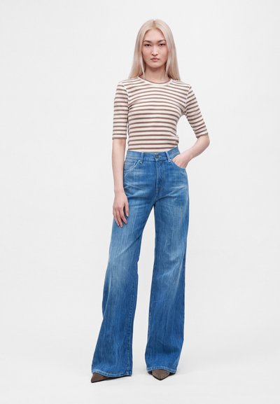 Femme aux longs cheveux blonds debout portant un haut rayé marron et blanc, un jean large bleu et des chaussures pointues marron sur fond blanc.