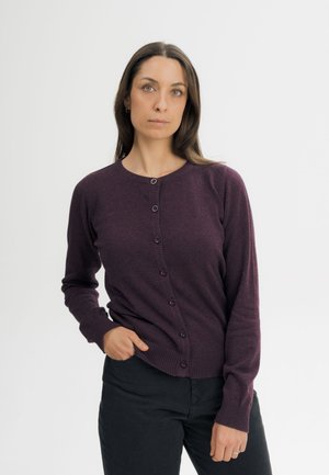 Vrouw met lang donker haar die een donkerpaarse gebreide cardigan met knopen en een zwarte broek draagt, staand tegen een effen lichte achtergrond.