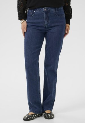 Vrouw in donkerblauwe rechte jeans, zwart longsleeve kanttop en zwarte flats met zilveren studs, staande met de handen langs het lichaam.