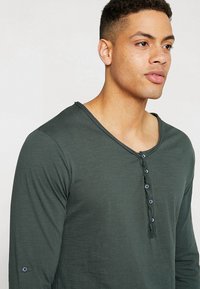 Dunkelgrünes Langarm-Henley-Shirt mit V-Ausschnitt, fünf Knöpfen und subtiler Textur. Mit kleinem Logo am Ärmel.
