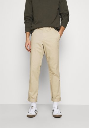 Beige cotton chinos with a straight fit, featuring rolled cuffs and side pockets, paired with white sneakers and an olive green sweatshirt.