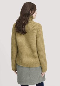 Senfgelbes Strickpullover mit hohem Kragen und texturiertem, geripptem Design. Getragen über einem grauen Cordrock mit geradem Schnitt.