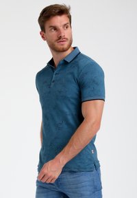 Polo teal avec un motif de feuilles subtil, col côtelé, patte de boutonnage à trois boutons et poignets contrastés. Porté avec un jean bleu clair.