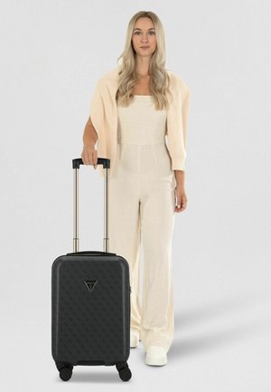 Frau in hellbeigem Outfit hält den Griff eines schwarzen Rollkoffers mit Muster und vier Rollen vor einfachem Hintergrund.