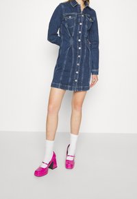 Džínová košilová šaty s dlouhými rukávy, knoflíkovým zapínáním a dvěma náprsními kapsami. V kombinaci s lesklými růžovými botami na blokovém podpatku a bílými ponožkami.