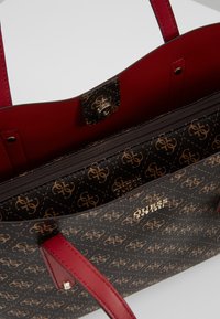 Zwarte handtas met een bruin logopatroon, rode voering en leren bandjes. Voorzien van een zak met rits en metalen accenten.