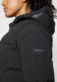 Čierna puffrová bunda s kapucňou, vyrobená z textúrovanej tkaniny, s prešívaným dizajnom a štítkom na rukáve s nápisom "ICEPEAK since 1996."