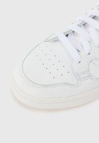 Valge nahast sneaker tekstureeritud pinnaga, auklik varvas, kontrastne õmblus ja valged sidemed. Ümar varvas ja veidi tõstetud tallad.