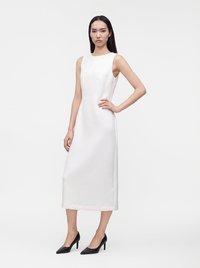 Femme aux longs cheveux noirs portant une robe midi blanche sans manches et des talons hauts noirs, debout avec une main sur la hanche sur un fond blanc.