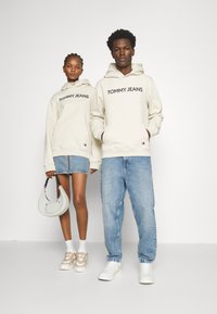 Sudaderas beige con "TOMMY JEANS" impresas en negro, combinadas con pantalones cortos de mezclilla azules y jeans claros. Un bolso ligero y zapatillas completan el look.