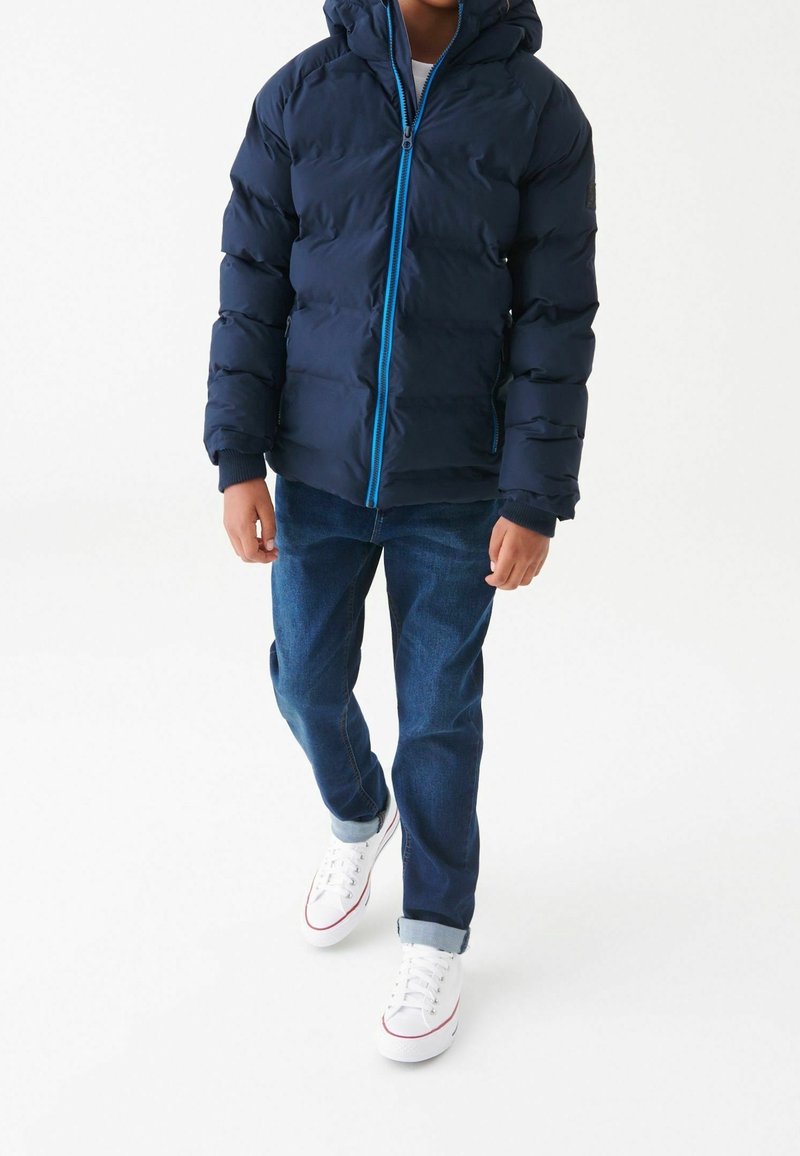 Marineblå puffjakke med hætte, blå lynlås, elastiske manchetter, parret med blå jeans og hvide sneakers med røde detaljer.