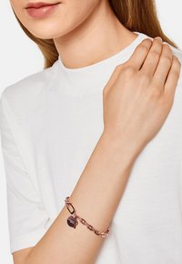 Bracelet en chaîne en or rose avec un fermoir, ornée d'un charm en forme de cœur. Le design comprend des maillons entrelacés et une texture lisse.