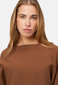 Pull en maille côtelée marron avec un col large, une texture douce et une coupe ample. Le modèle présente des cheveux blonds mi-longs et peu d'accessoires.