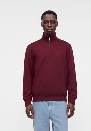 Mann trägt einen burgunderfarbenen Viertel-Reißverschluss-Pullover und hellblaue Jeans, steht vor einem schlichten hellen Hintergrund.