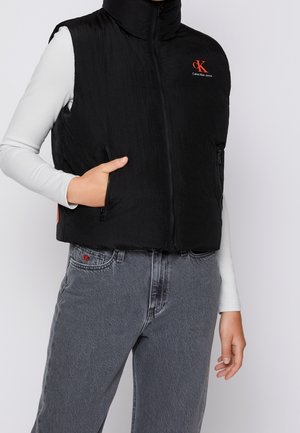 Person iført sort zip-up Calvin Klein polstret vest over hvid langærmet top og grå jeans med rødt CK-logo på lommen, hånden i vestens lomme.