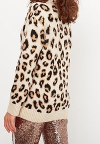 Maglione a maglia color crema con stampa leopardata nera e marrone, dotato di orlo e maniche a coste. Indossato con pantaloni lucidi di paillettes.