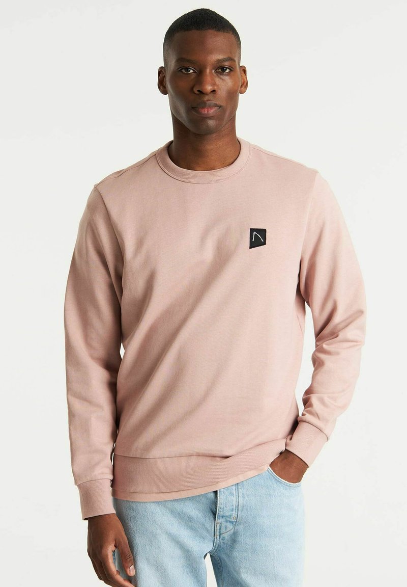 Sudadera de color rosa claro, hecha de algodón, con cuello redondo, mangas largas, puños de canalé y un pequeño parche negro con el logo en el pecho.