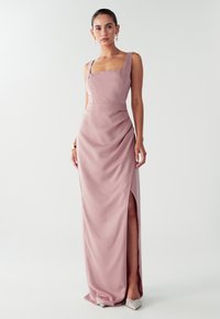 Moteris vilkinti grindimis ilgo mėtų atspalvio satino suknelę su plataus petnešomis, šoniniu įpjovimu ir sidabriniais dekoruotais suknelės bateliais, stovinti prieš baltą foną.