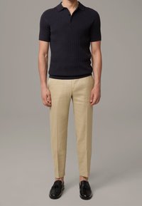Polo en maille marine à manches courtes, associé à un pantalon tailleur beige clair et des mocassins à lacets noirs. Textures lisses et design ajusté.