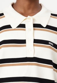 Stripet polo-skjorte i kremfarge, svart og brun. Har kragehals og fire knapper, med en liten logo brodert på brystet.