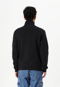 Zwarte sweatshirt met lange mouwen en hoge kraag, gemaakt van zacht, textuurstof. Heeft ribbelmanchetten en -zoom. G gedragen met lichtblauwe cargobroek.