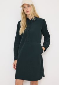 Blonde Frau, die eine beigefarbene Mütze und ein langärmliges schwarzes Knopfkleid mit Taschen trägt, steht vor einem weißen Hintergrund.