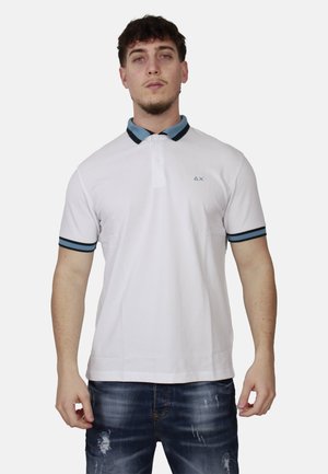 Uomo che indossa una polo bianca a maniche corte con colletto e bordi delle maniche azzurri e neri, abbinata a jeans blu strappati, in piedi di fronte a chi guarda.