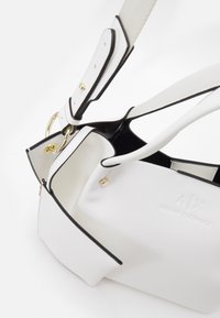 Bílá texturovaná kabelka s černými detaily, která má zlatý kroužkový hardwar, nastavitelný popruh a značení s raženým logem.