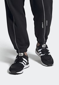 Svarta sportskor med vita tre-stripsdetaljer, med material av mocka och mesh, rund tå och gummisula. Matcher med svarta joggers.