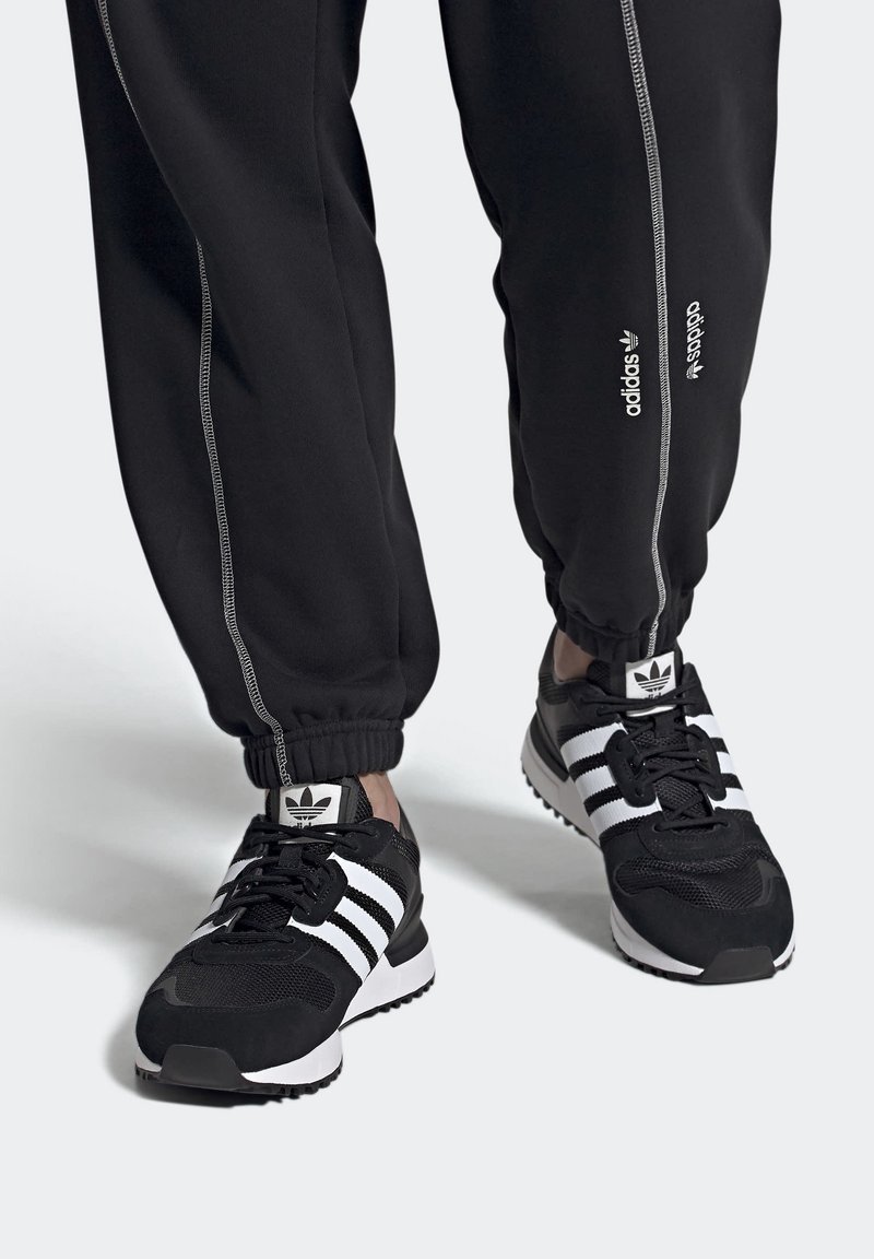 Svarta sportskor med vita tre-stripsdetaljer, med material av mocka och mesh, rund tå och gummisula. Matcher med svarta joggers.