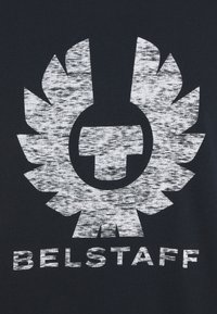 Schwarzer Stoff mit einem auffälligen weißen Logo, das ein rundes Emblem in der Mitte zeigt, umgeben von stilisierten flügelförmigen Formen. Der Text "BELSTAFF" befindet sich darunter.