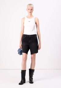 Un débardeur blanc avec le logo "GAP", associé à un short en denim noir taille haute. Le mannequin porte des bottes noires jusqu'aux genoux et tient un objet bleu.