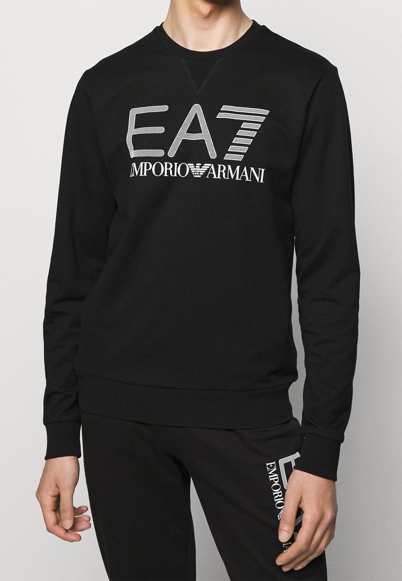 Kvinna i svart långärmad sweatshirt och byxor med den vita "EA7 Emporio Armani" logotypen tryckt på bröstet och låret.