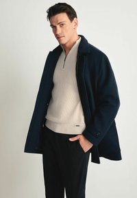 Granatowy wełniany płaszcz z wyciętym kołnierzem, wytłaczany kremowy sweter i czarne spodnie. Płaszcz ma ozdobne guziki i luźny krój.